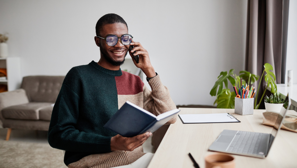 Portrait of young African-American man wearing glasses while working from home and speaking by smartphone,