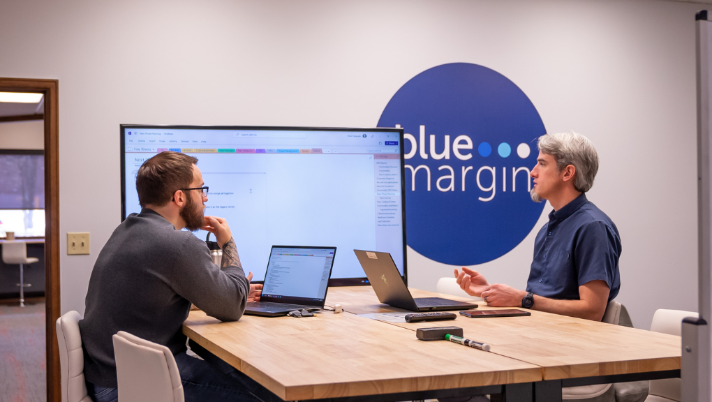 Two co-workers on their computer collaborating with each other. There is a Blue Margin logo in the background.