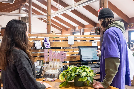 Cashier wearing a purple shirt and black hat checking out a customer in a greenhouse.