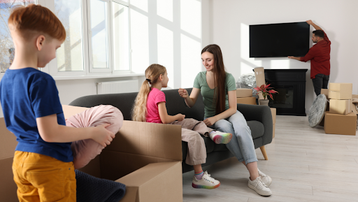 woman in green shirt sitting on black couch with little girl in pink shirt with moving boxes around them. a tv hangs on the wall behind them.