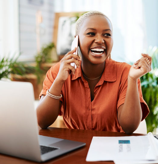 A woman in an orange shirt on a cell phone laughing while working on a computer