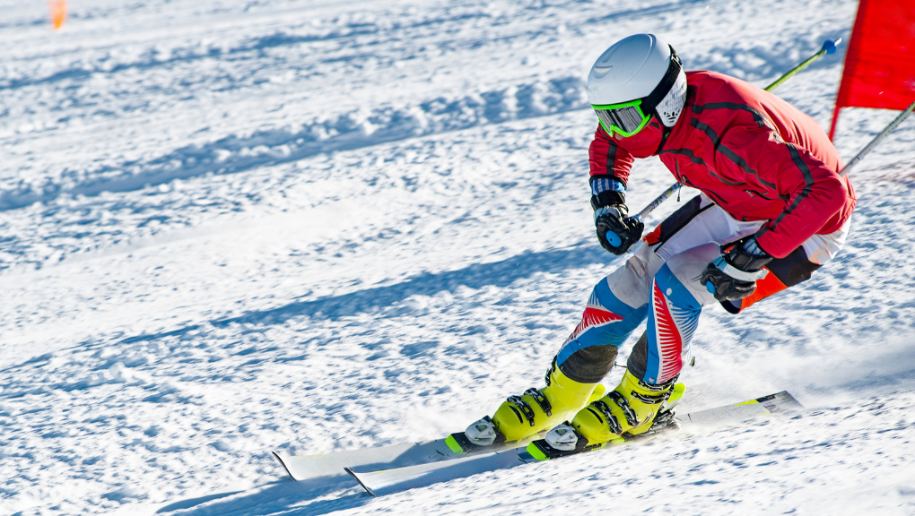 Skier in a red jacket and helmet races downhill on a snowy slope, leaning into a turn near a red course flag.
