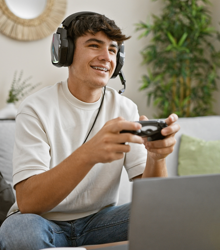 Cheerful, young hispanic teenager student, gamepad in hand, engrossed in online gaming, lounging on living room sofa, making his apartment a lively gaming house.