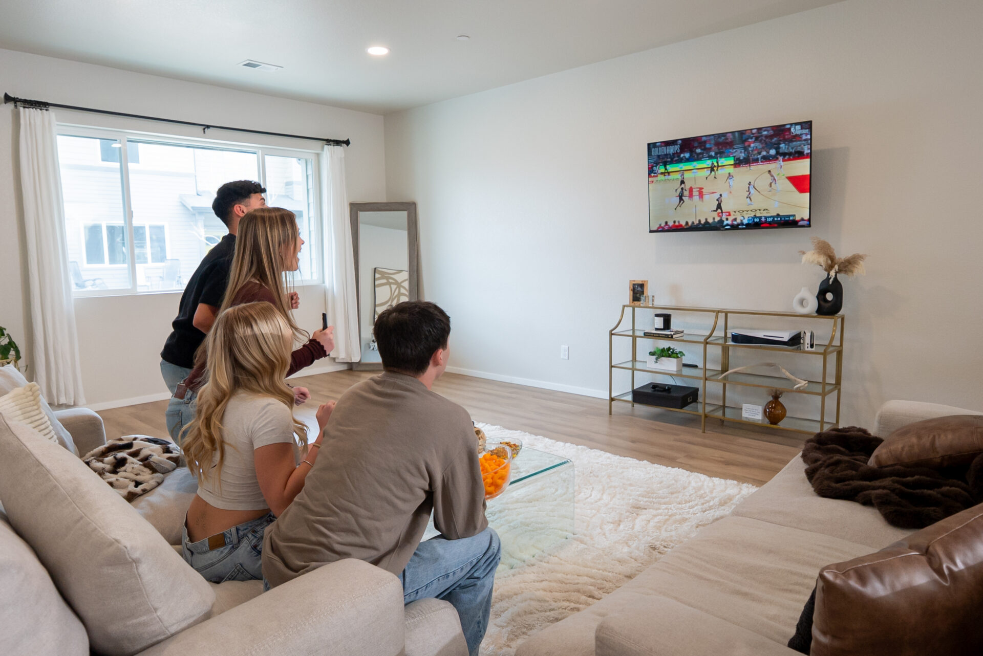 Four young adults in a living room watch a basketball game on a wall-mounted TV. Two are standing, two are seated on a couch, all focused on the screen. The room is bright with modern decor.