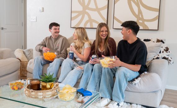 cx-student-pricing-verification Four young adults sit together on a living room couch, laughing and talking while holding bowls of snacks and drinks. A coffee table filled with chips, popcorn, and candy sits in front of them, creating a relaxed and social gathering atmosphere.