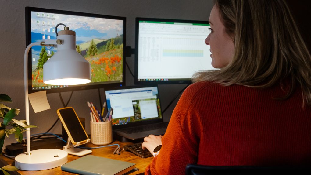 Employee working at a desk with dual monitors and a laptop, reviewing spreadsheets and business data in a home office. The workspace includes a desk lamp, mobile phone, notebook, and office supplies, creating a focused remote work environment.