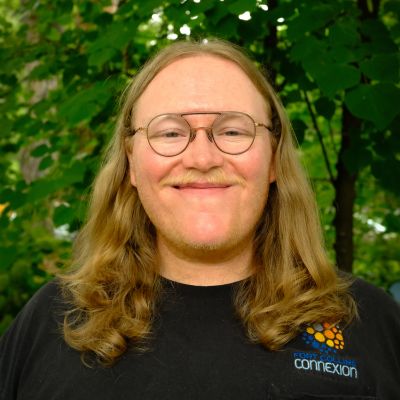 A person with long light brown hair and glasses smiles outdoors, wearing a black shirt that says “connexion” with an orange and blue logo. Green leafy trees fill the background.