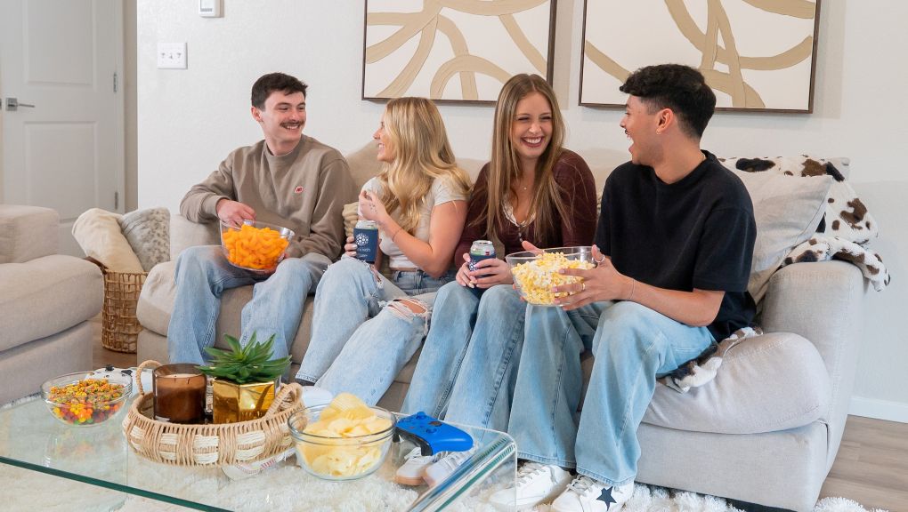 Four young adults sit together on a living room couch, laughing and sharing snacks and drinks, with a video game controller on the coffee table.