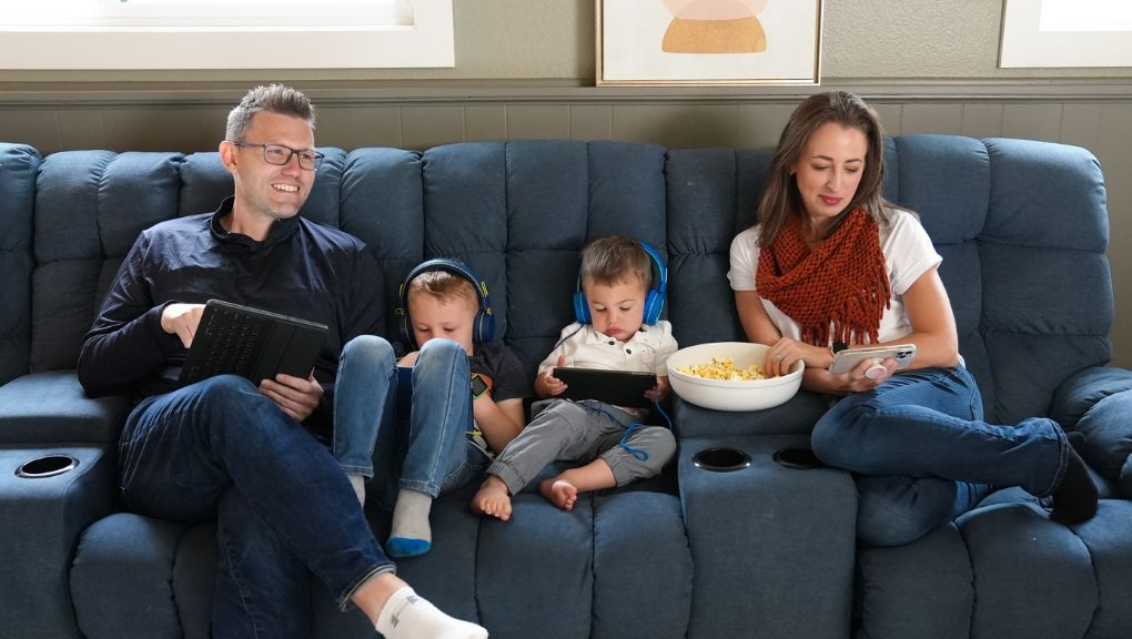 A family of four sits together on a blue couch, with the parents using a tablet and smartphone while two young children wear headphones and use mobile devices.